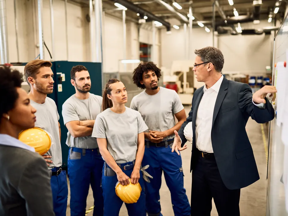 Um consultor de terno apresenta um plano num quadro branco para um grupo de operadores de fábrica.
